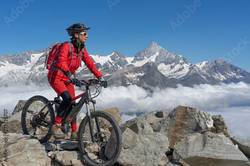 Fototapeta na drzwi i meble aktywna starsza kobieta, jadąca elektrycznym rowerem górskim Gornergrat w Zermatt, Wallis, Szwajcaria. W tle Weisshorn, Zinalrothorn i Obergabelhorn