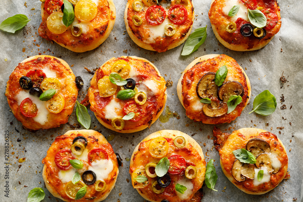 Mini vegetarian pizzas with the addition various kinds of vegetables , mozzarella cheese and fresh basil on a on white baking paper, close-up, top view. - obrazy, fototapety, plakaty 