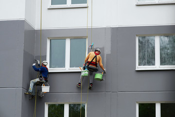 Worker hanging on rope and paints building wall with roller. Painter hanging on cable with paint buckets, industrial climber repairing house facade. Industrial alpinist and climbing. Rigging equipment