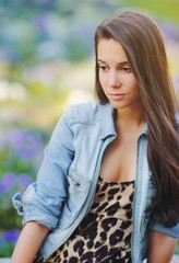 Stylish long hair brunette woman in a leopard animal print dress in denim jacket, street fashion concept. Fashionable hipster girl sitting on summer bench with flower background. Trendy boho style.