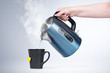 Female hand pours hot water from an electric kettle into a black mug, on light background.