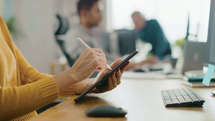 Poster - Anonymous Young Woman Sitting at Her Desk She's Drawing, Writing and Using Pen with Digital Tablet Computer. Focus on Hands with Pen. Bright Office where Diverse Team of Young Professionals Work 