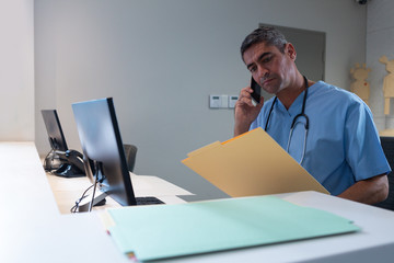 Male surgeon talking on mobile phone while looking at medical report at desk