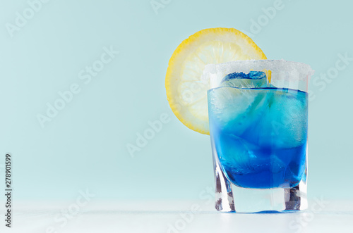 Exotic cold shot glass cocktail with yellow lemon slice, blue curacao, ice cubes, salt rim on soft light mint color background and white wood board Fotobehang