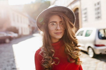 Wall Mural - Calm long-haired girl posing on the street in sunny autumn day. Catching curly woman in glasses enjoying sunshine during outdoor photoshoot.