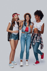 Poster - Full length of three attractive young women drinking refreshing cocktail and smiling while standing against grey background