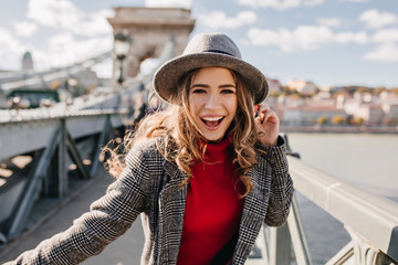 Wall Mural - winsome curly girl in elegant gray hat posing on bridge in sunny day. fashionable female tourist spe