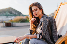 Serious Blue-eyed Lady Chilling In Outdoor Restaurant On Mountain Background. Portrait Of Brown-haired Stylish Woman In Gray Coat Posing In Recliner In Cafe.