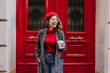© Look! - Glad white female model in long gray jacket looking away with smile standing near red door. Outdoor photo of beautiful caucasian woman in sweater drinks coffee in front of cafe and laughing.