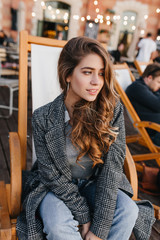 Wall Mural - Pensive blue-eyed girl with pale skin sitting in recliner in outdoor cafe and looking away. Shy brunette caucasian woman in warm coat waiting someone in cold day.