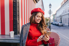 Inspired Woman With Pleased Face Expression Texting Message While Drinking Coffee Near Outdoor Cafe. Portrait Of Curious Girl In Red Beret Checking Mobile Mail On The Street.