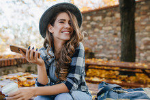 Pretty Laughing Girl With Smartphone Has A Good Time In Autumn Weekend. Outdoor Portrait Of Lovable Trendy Lady With Brown Hair Wears Hat In October Day.