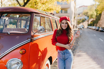 Adorable female model with black manicure posing on the road with charming smile in warm autumn day. Lovely white girl in beret standing near orange bus and waiting for driver.