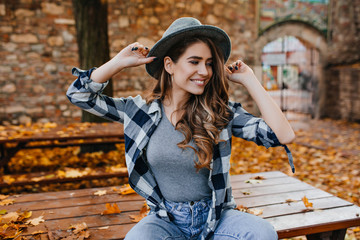 Magnificent pale girl in trendy hat sitting on table in park and smiling. Adorable young woman with curly hairstyle playfully posing, enjoying autumn weekend.