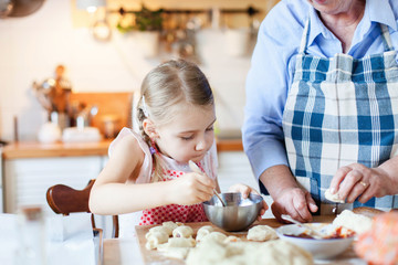 Family is cooking in cozy kitchen at home. Grandmother is teaching little girl. Retired woman and child make pastry dough together. Cute kid is helping to prepare meal. Lifestyle authentic moments.