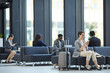© Seventyfour - Business people wasting time in airport: cheerful attractive woman sitting on sofa and reading interesting articles on internet