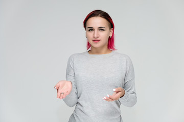 Portrait to the waist of a pretty girl with red hair on a white background in a gray sweater. Standing right in front of the camera in a studio with emotions, talking, showing hands, smiling