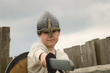 A Boy In A Historical Costume Of Medieval War With Helmet And Shield Put Forward A Wooden Sword Standing In The Middle Of A Wooden Fortress Outdoors In Summer