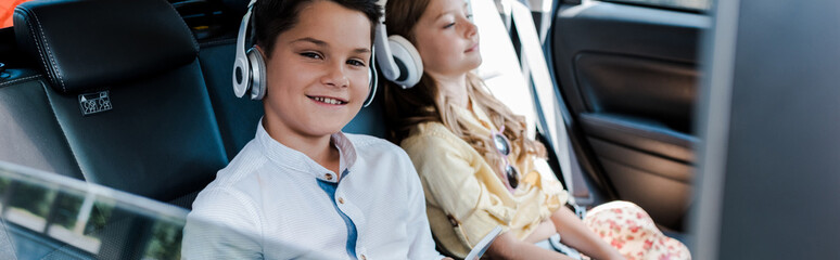 Wall Mural - panoramic shot of boy listening music in headphones near sister in car