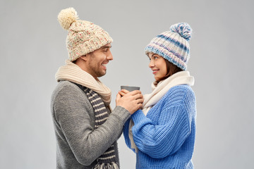 Wall Mural - people, christmas and love concept - happy romantic couple in knitted hats and scarves holding one cup over grey background