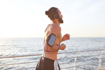 Photo of young handsome sporty bearded guy, listen new cool music on headphones and running at the seaside. Enjoy the morning and sunset.