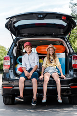 Wall Mural - happy kid in cap sitting near sister in straw hat and looking at camera