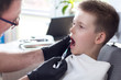 Dentysta w czarnych gumowych rękawiczkach trzyma w dłoni strzykawkę z igłą i robi zastrzyk chłopcu siedzącemu na fotelu dentystycznym z otwartymi ustami.