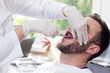 Biały mężczyzna siedzi na fotelu dentystycznym z szeroko otwartymi ustami. Dłonie stomatologa w białych rękawiczkach chwytają zęba kleszczami.