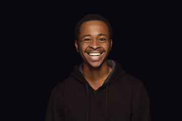 Close up portrait of young african-american man isolated on black studio background. Real emotions of male model. Standing and looks happy, smiling. Facial expression, human nature and emotions