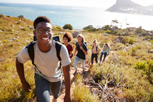Millennial African American Man Leading Friends Hiking Single File Uphill On A Path By The Coast
