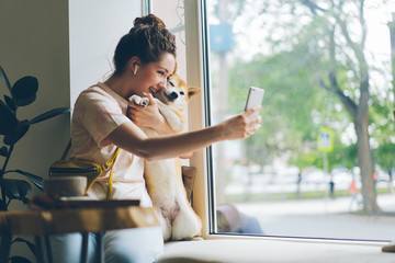 Cheerful lady taking selfie with doggy in cafe using smartphone camera sitting on window sill having fun with cute animal. People and technology concept.