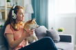 Beautiful woman in headphones is listening to music and petting pedigree dog sitting on couch enjoying song holding smartphone. Happiness, animals and youth culture concept.