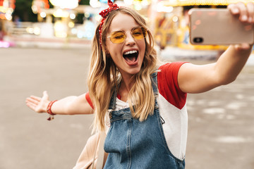 Poster - Image of joyful blonde woman taking selfie photo on cellphone in front of colorful carousel at amusement park