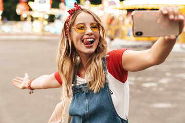 Sticker - Image of cheerful blonde woman taking selfie photo on cellphone in front of colorful carousel at amusement park