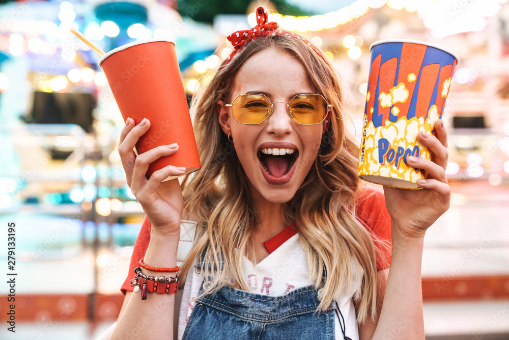 Image of joyful charming woman holding popcorn and soda paper cup while walking in amusement park - obrazy, fototapety, plakaty 