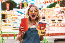 Image Of Excited Charming Woman Holding Popcorn And Soda Paper Cup While Walking In Amusement Park