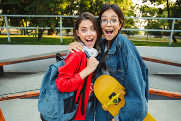 Poster - Image of two excited girls smiling and hugging together while holding skateboard in skate park