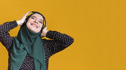 Happy muslim woman listening songs on headphone against yellow surface