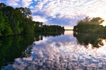 Wall Mural - River with reflections from clouds and a tower and forest on the left side and forest and sun on the right side