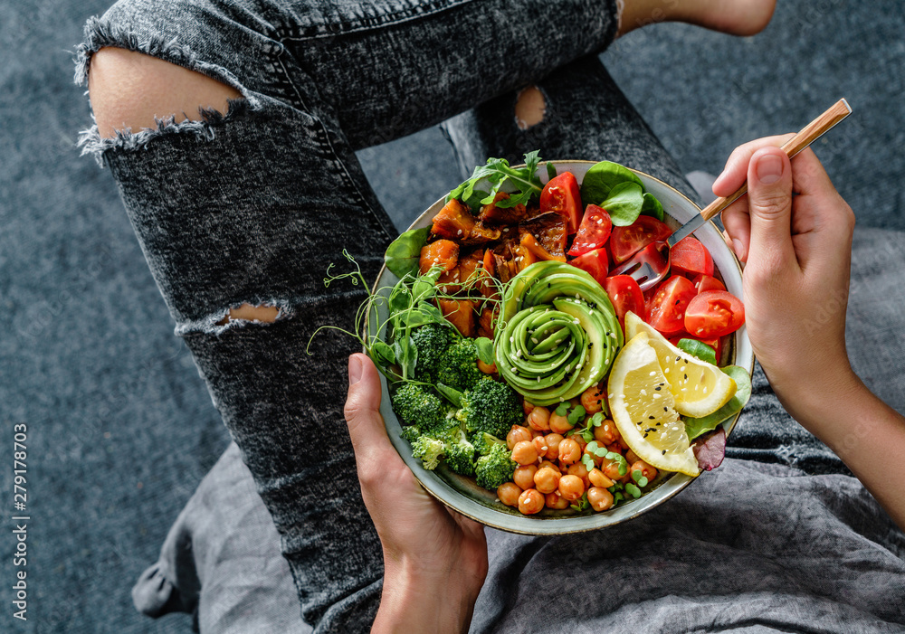 Woman in jeans holding Buddha bowl with salad, baked sweet potatoes, chickpeas, broccoli, greens, avocado, sprouts in hands. Healthy vegan food, clean eating, dieting, top view - obrazy, fototapety, plakaty 