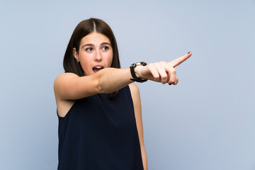 Wall Mural - Young woman over isolated blue wall pointing away