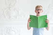 © stock28studio - A little schoolboy in a glasses is reading aloud text from his book on background of white wall