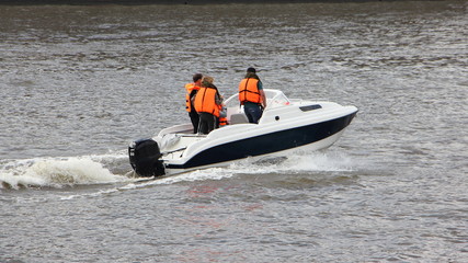 Wall Mural - White plastic cabin motor boat with passengers in orange life jackets quickly floats on the river on a summer day