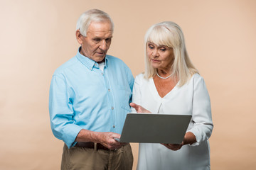 Wall Mural - retired woman gesturing while looking at laptop near senior husband isolated on beige