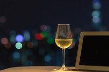 A Glass With White Wine Puts On Sofa With Laptop Computer With Colorful Bokeh Light From City.