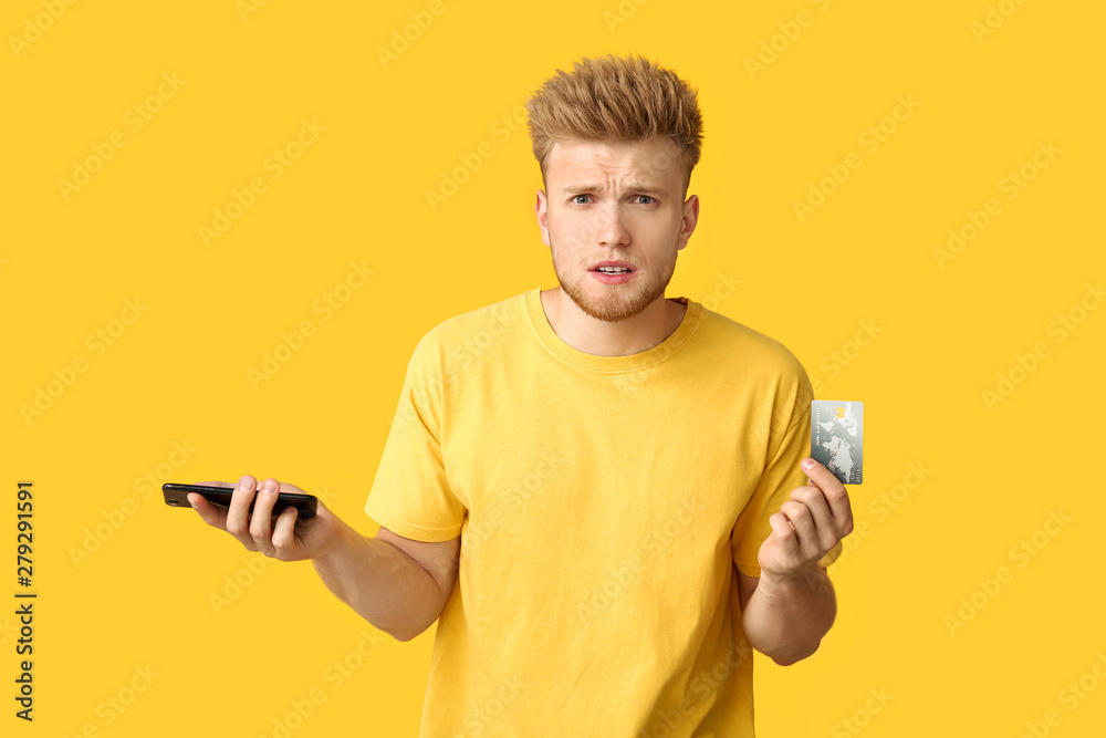 Stressed young man with credit card and mobile phone on color background