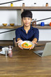 © Thanadol - Young asian man showing breakfast with coffee and laptop on wooden table in shelf background of kitchen room