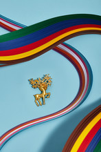 Closeup Shot Of Golden Brooch In The View Of Red Deer, Isolated Asymmetrically On Light Blue Background In The Midst Of Multicolored Wavy Placed Ribbons. Voguish Fashion Item. Fashionable Adornment.