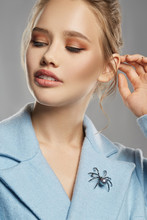 Portrait Of Lady With Tied Back Fair Hair, Wearing Sky Blue Coat With Massive Brooch In View Of Gray Spider With Pearly Insert On Lapel. The Girl Is Turning Head To Side Against The Gray Background.