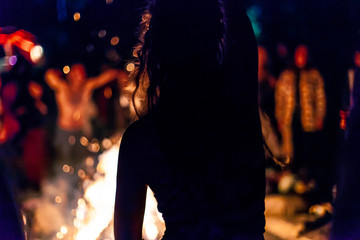 Fusion of cultural & modern music event. A silhouetted girl is seen from behind as she dances by a campfire during a music festival, blurry people and warm glow provides backlight by night